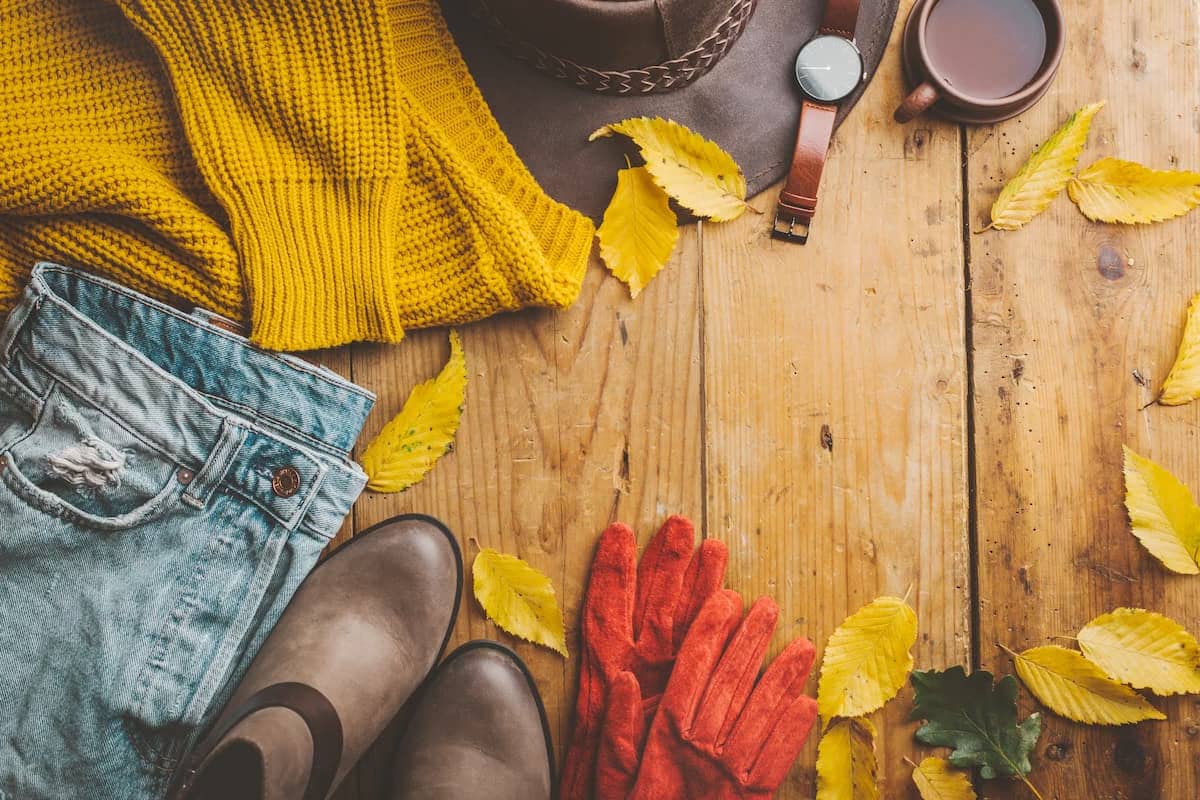 A flat lay of pre-loved women's fall clothing and accessories on a rustic wooden surface, featuring a mustard yellow knit sweater, light wash denim jeans, brown ankle boots, red knit gloves, a brown fedora hat, and a mug of tea or coffee. Scattered yellow leaves add an autumnal touch.