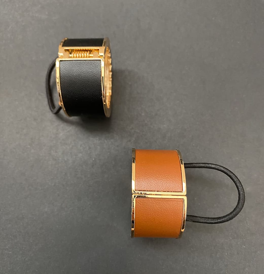 two leather and metal ponytail covers with elastic, shown in black and tan leeather