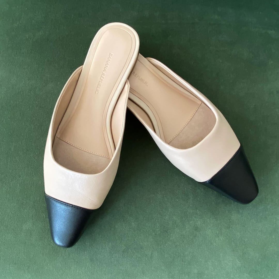 A pair of new Banana Republic beige ballet mules with black accents, displayed on a green textured background. The backless flats feature a pointed toe and a sleek two-tone design, inspired by Chanel.